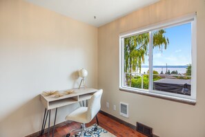 A room with a desk, chair, and a large window with a view of a body of water and greenery.