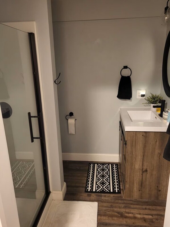 Un baño con mampara de vidrio, lavamanos blanco, tocador de madera, toalla negra y alfombra estampada.