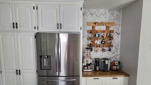 Una cocina con gabinetes blancos, un refrigerador de acero inoxidable, una estación de café con tazas y un salpicadero con diseño de espiga.