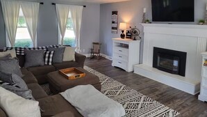 Una sala de estar con un sofá marrón, una chimenea blanca y un televisor grande montado en la pared.