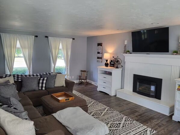 Een woonkamer met een bruine bank, een witte schoorsteen en een grote televisie aan de muur.
