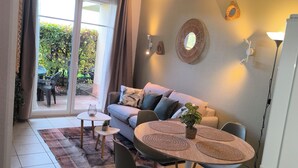 A modern living room with a sofa, a dining table with chairs, a ceiling light, and a view through a glass door to an outdoor area.