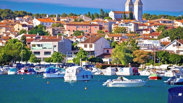 Une ville côtière qui se distingue par son église et son clocher imposants, entourée de bâtiments résidentiels et d’un port de plaisance où sont amarrés des bateaux.
