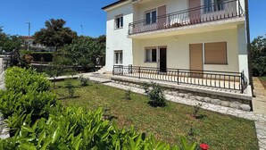 A two-story building with a balcony on the upper floor, a stone pathway leading to the entrance, and a well-maintained garden in the foreground. - Holiday apartment mit Klimaanlage und Terrasse (Krk)
