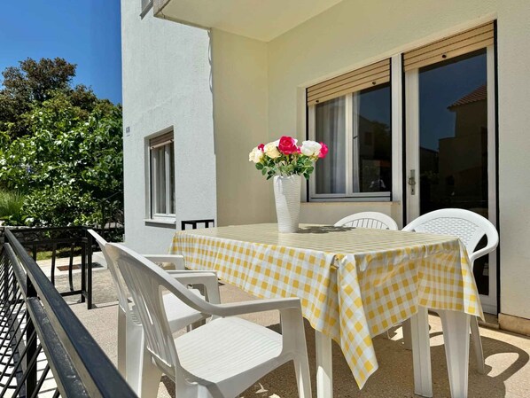 Une terrasse extérieure avec une table recouverte d’une nappe à carreaux jaunes et blancs, entourée de chaises en plastique blanches. Un vase blanc contenant des fleurs roses et blanches est posé sur la table.