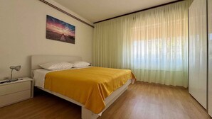 A bedroom with a bed, a nightstand, a lamp, a painting, and a window with sheer curtains. - Holiday apartment mit Klimaanlage und Terrasse (Krk)