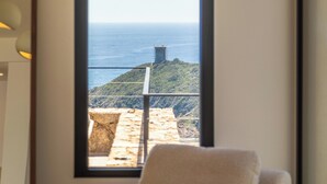 Une pièce dotée d’une grande fenêtre donnant sur un paysage côtier, d’un fauteuil confortable et d’une petite table sur laquelle est posé un chapeau.