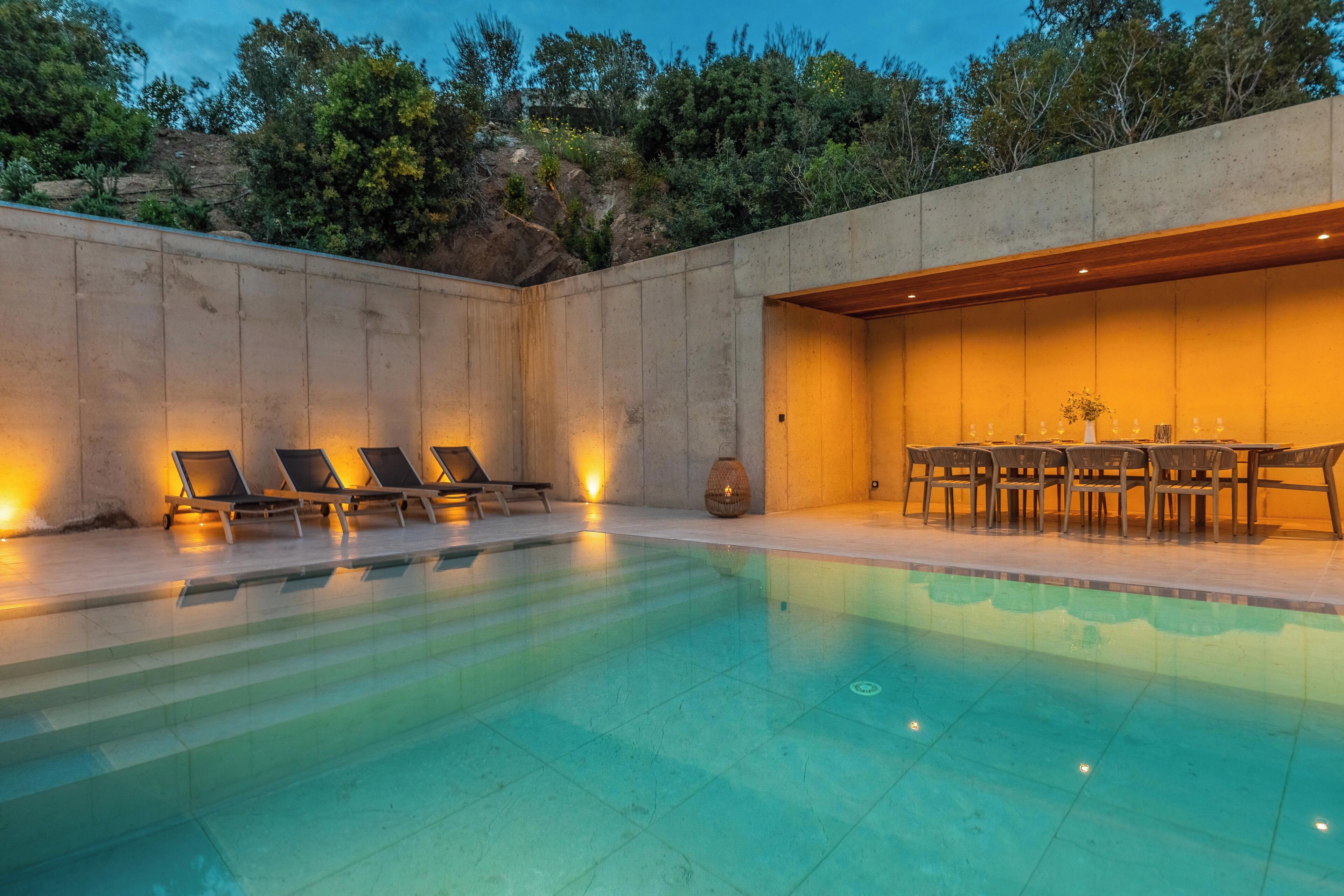 A swimming pool with lounge chairs and a dining area, surrounded by a concrete wall and greenery.