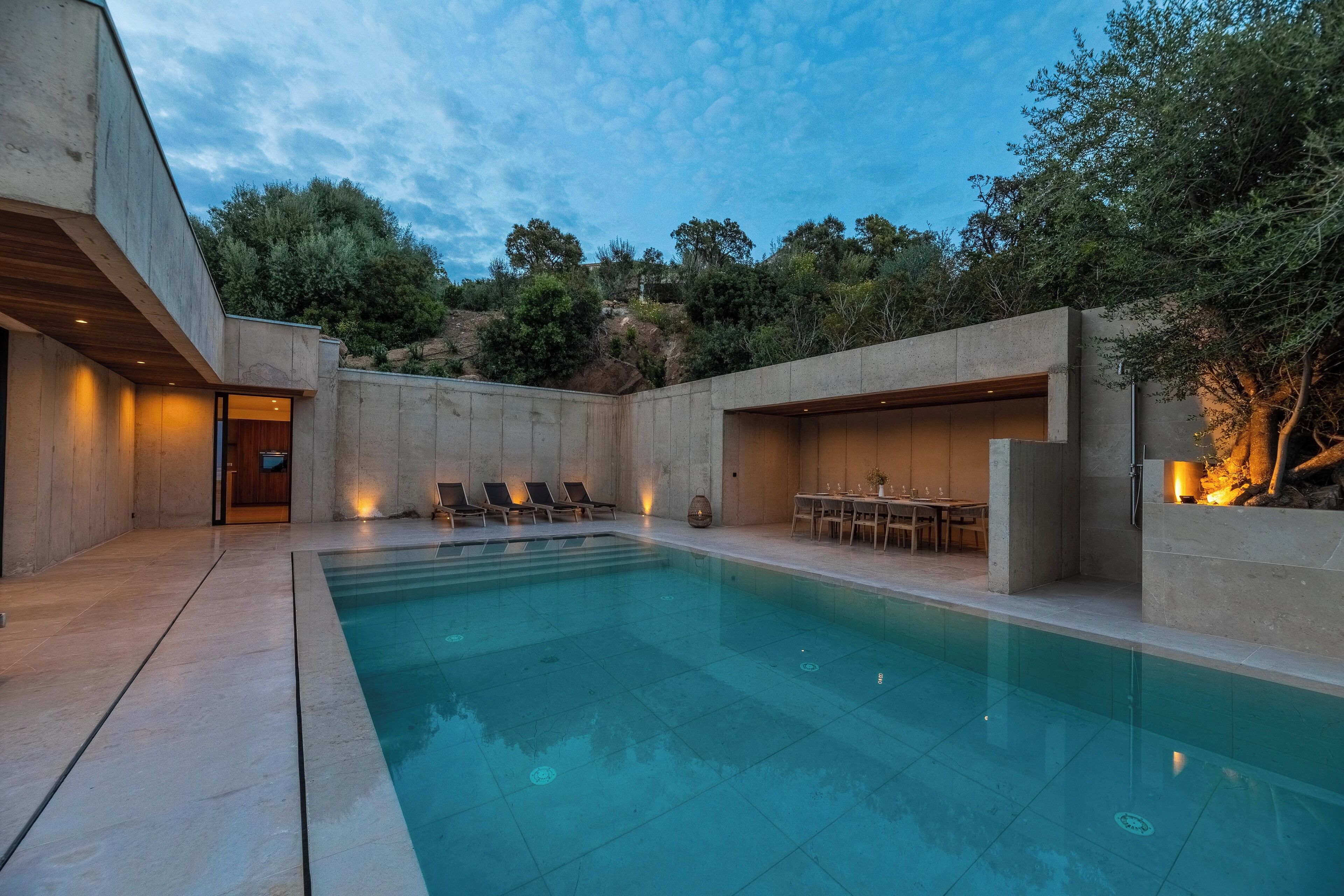 A modern outdoor pool area with a concrete pool, lounge chairs, and a dining table.