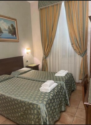 A hotel room with two beds, a nightstand, a lamp, a painting, and curtains.