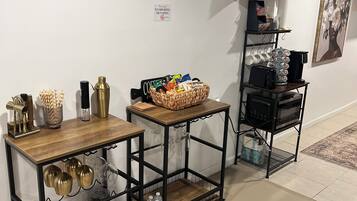 A room with a wooden table, a metal cart with glassware, and a wall-mounted shelf with cups and a picture.