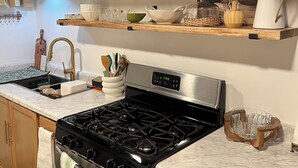 A kitchen with a gas stove, oven, wooden cabinets, marble countertops, and wooden shelves holding kitchenware.