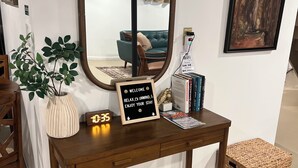 A wooden console table with a mirror, a digital clock, a welcome sign, books, and a framed painting.