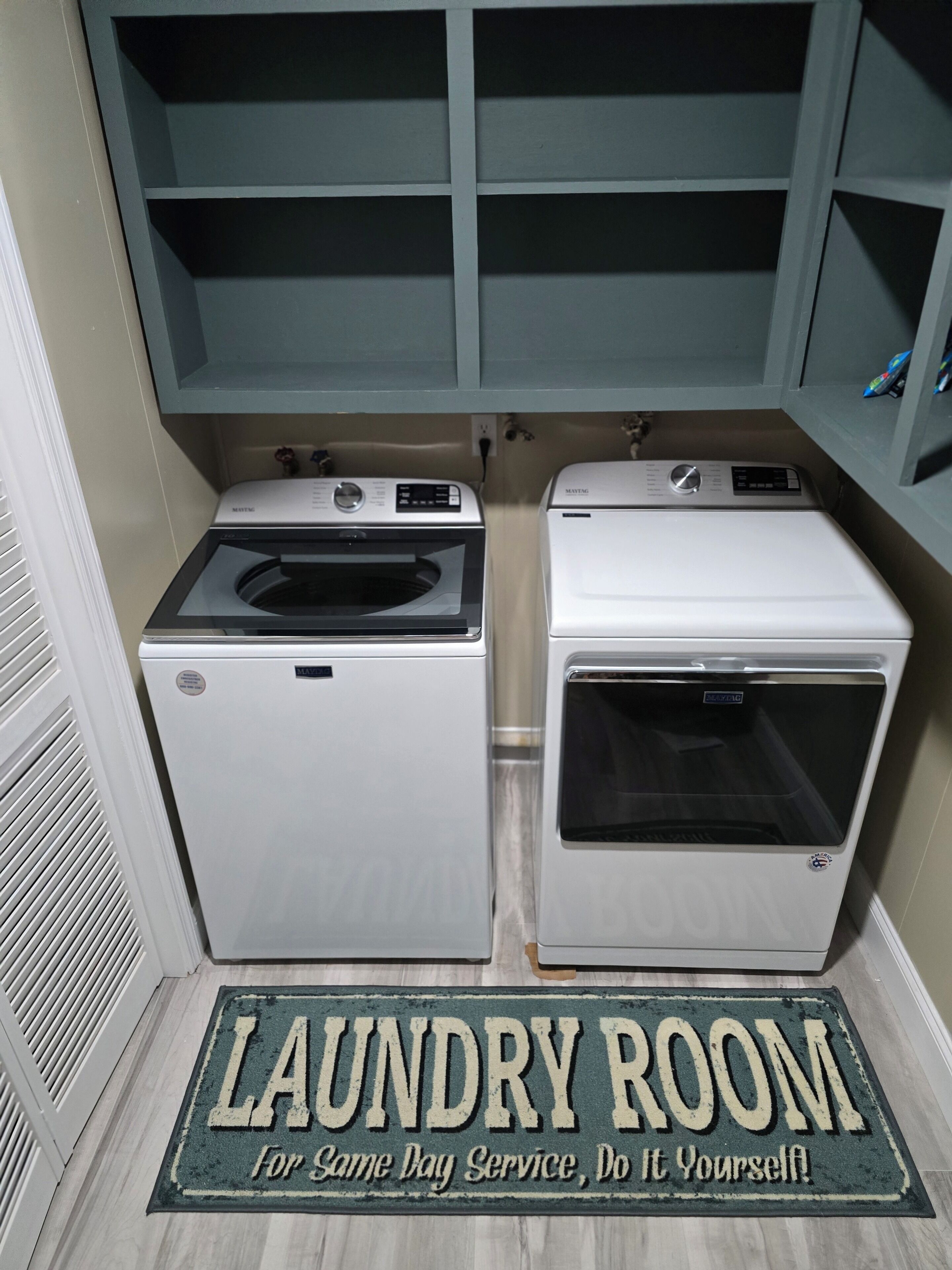 Une buanderie équipée de deux machines à laver et d’un sèche-linge, un paillasson avec l’inscription « BUANDERIE », et une étagère au-dessus des machines.