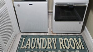 Un cuarto de lavandería con dos lavadoras y una secadora, un felpudo con el texto "LAUNDRY ROOM" y un estante sobre las máquinas.