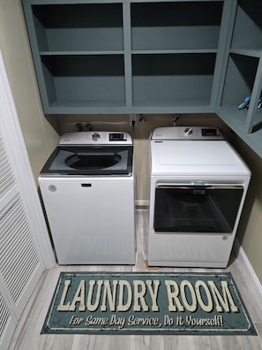 Un cuarto de lavandería con dos lavadoras y una secadora, un felpudo con el texto "LAUNDRY ROOM" y un estante sobre las máquinas.