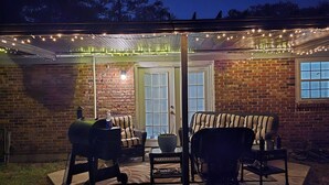 Un patio al aire libre con pared de ladrillo, dos sillas de mimbre, una parrilla y luces colgantes.