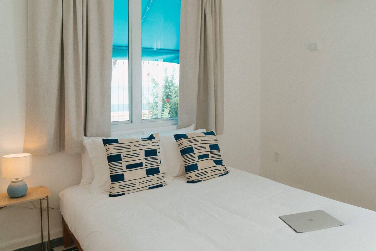 A neatly made bed with two patterned pillows, a laptop on the bed, and a lamp on a bedside table.
