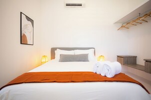 Una cama bien hecha con una colcha blanca, un edredón marrón y dos almohadas. En la pared, a la izquierda de la cama, hay una obra enmarcada.