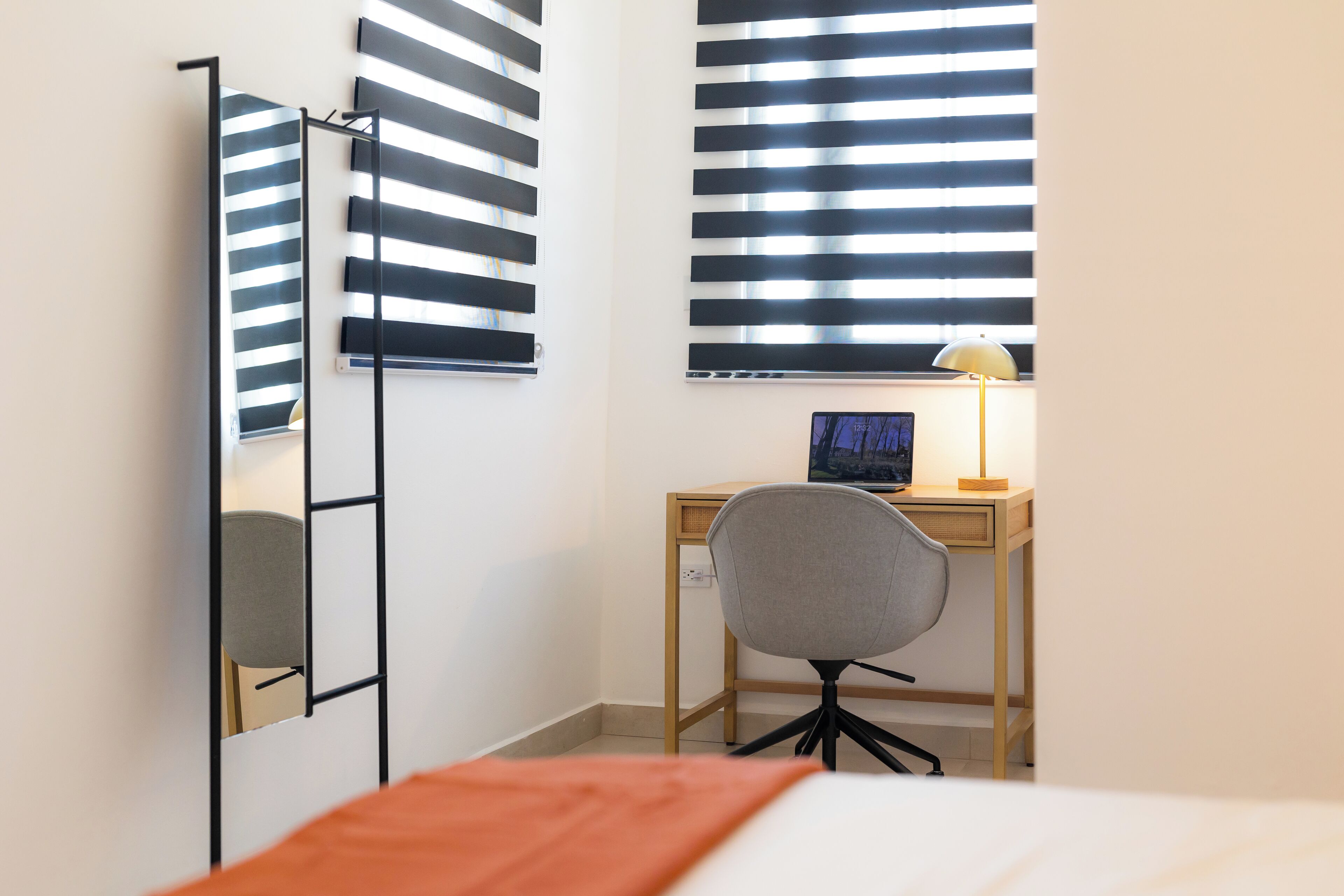 A bedroom with a bed, a desk with a chair, a lamp, and a window with blinds.