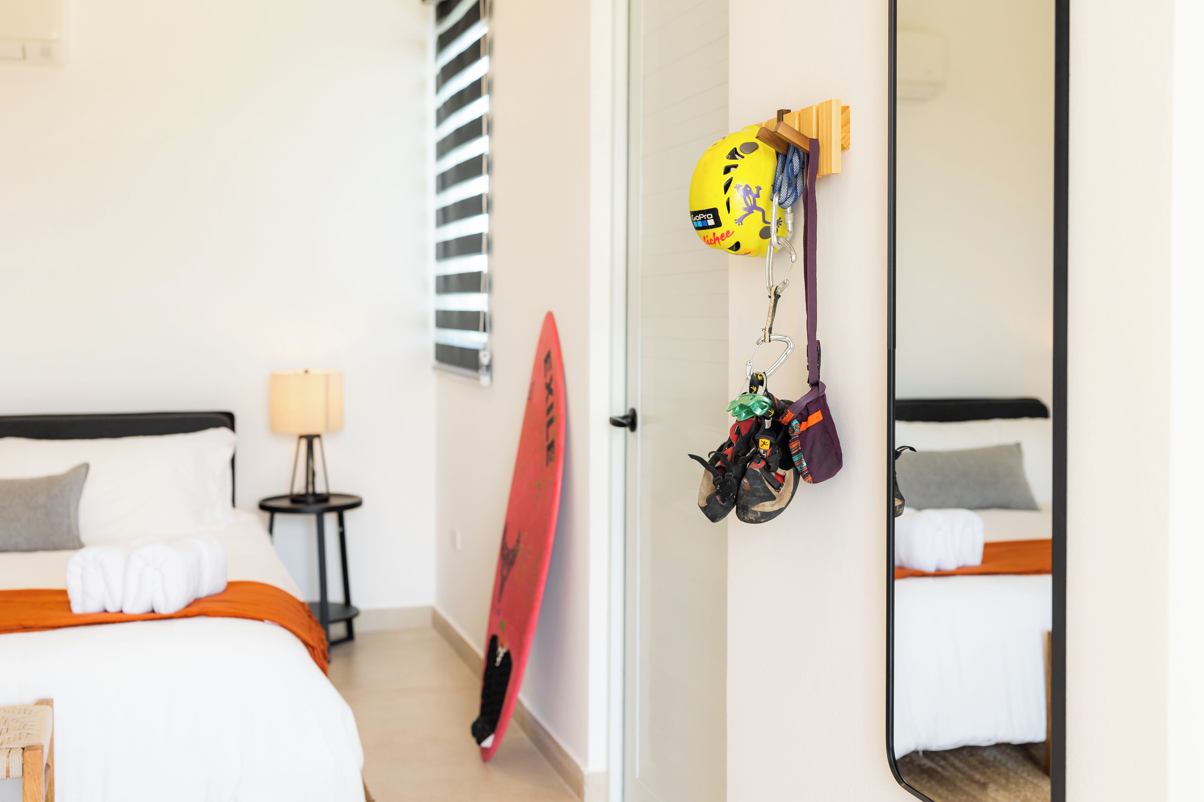 Ein Hotelzimmer mit zwei Betten, einem Nachttisch mit Lampe, einem an der Wand stehenden Surfbrett und einem Spiegel, der den Raum widerspiegelt.