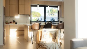 A modern kitchen with wooden cabinets, a large window, and a dining area with chairs. - Outsite Puerto Rico Aguadilla - Work, Connect, Explore (Aguacate)