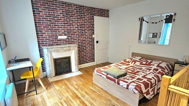 Une chambre avec un mur en briques, un lit avec une literie à motifs, un plancher en bois, un bureau avec une chaise et un miroir sur le mur.