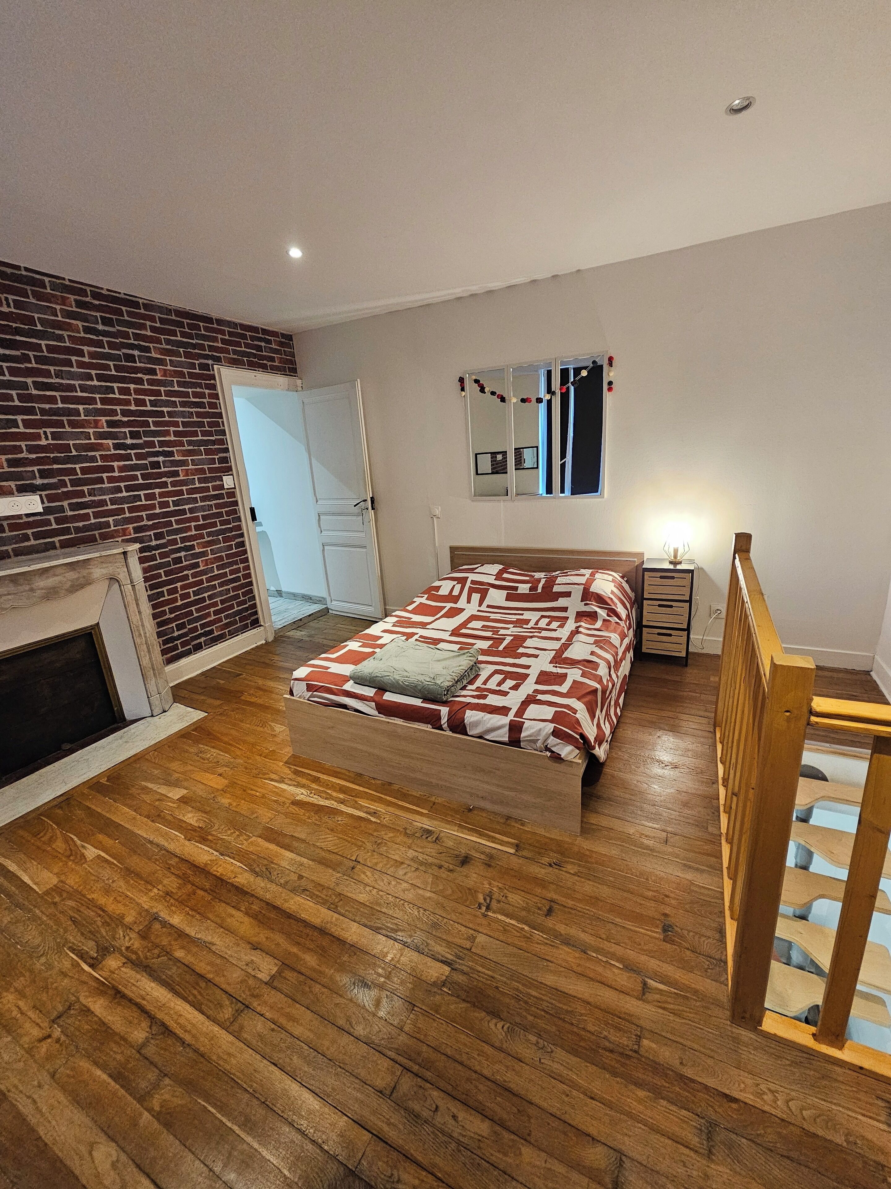 Ein Schlafzimmer mit Holzboden, einem Bett mit gemusterten Betttextilien, einer Ziegelwand, einer weißen Tür, einem kleinen Nachttisch mit Lampe und einem Spiegel an der Wand.