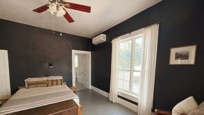 Ein Schlafzimmer mit einem Bett, einer Holzkommode, einem Deckenventilator, einem Fenster mit Vorhängen und einer Tür zu einem anderen Raum.