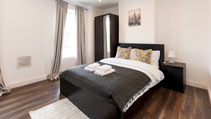 A modern bedroom with a dark wood floor, a large bed with patterned pillows, a nightstand with a lamp, and a window with curtains.