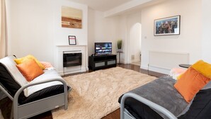 A modern living room with a fireplace, a flat-screen TV, and two beds with colorful pillows.