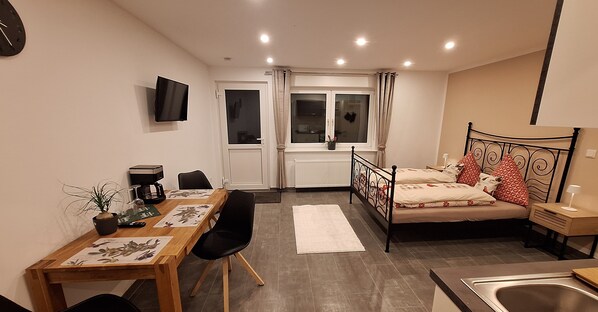A compact living space with a dining area, a bed with decorative pillows, a TV mounted on the wall, and a window with curtains. - Cozy, newly renovated vacation apartment "Merlin" in the heart of the Taunus! (Weilrod)