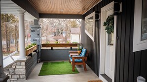 A covered porch with a wooden bench and a small table, a green artificial grass mat, and a stone planter with plants.