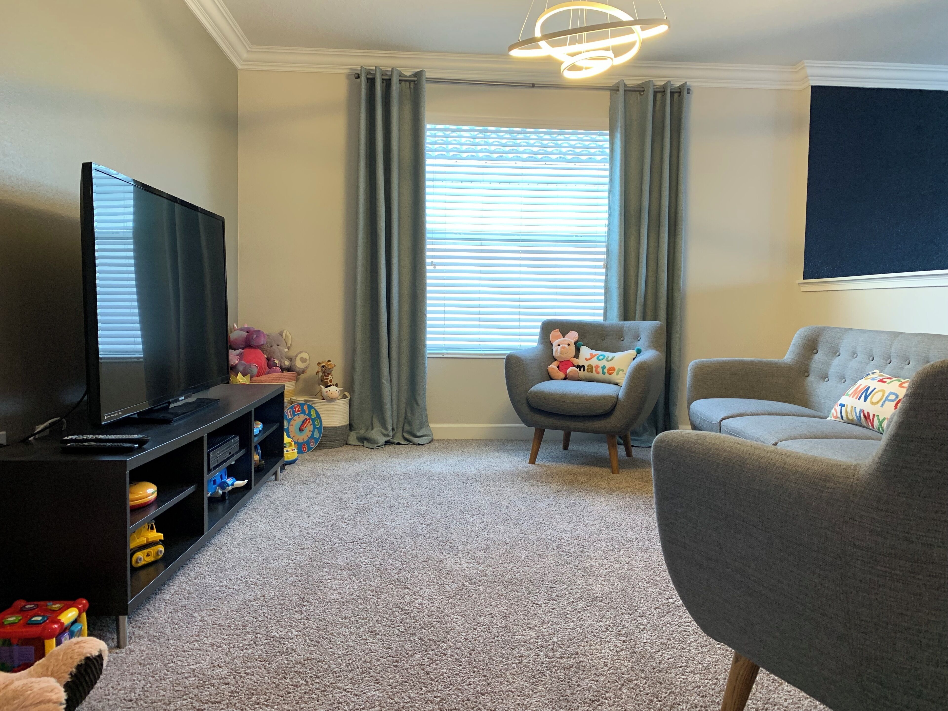 A living room with a television, a grey armchair, a sofa, and a window with blinds.