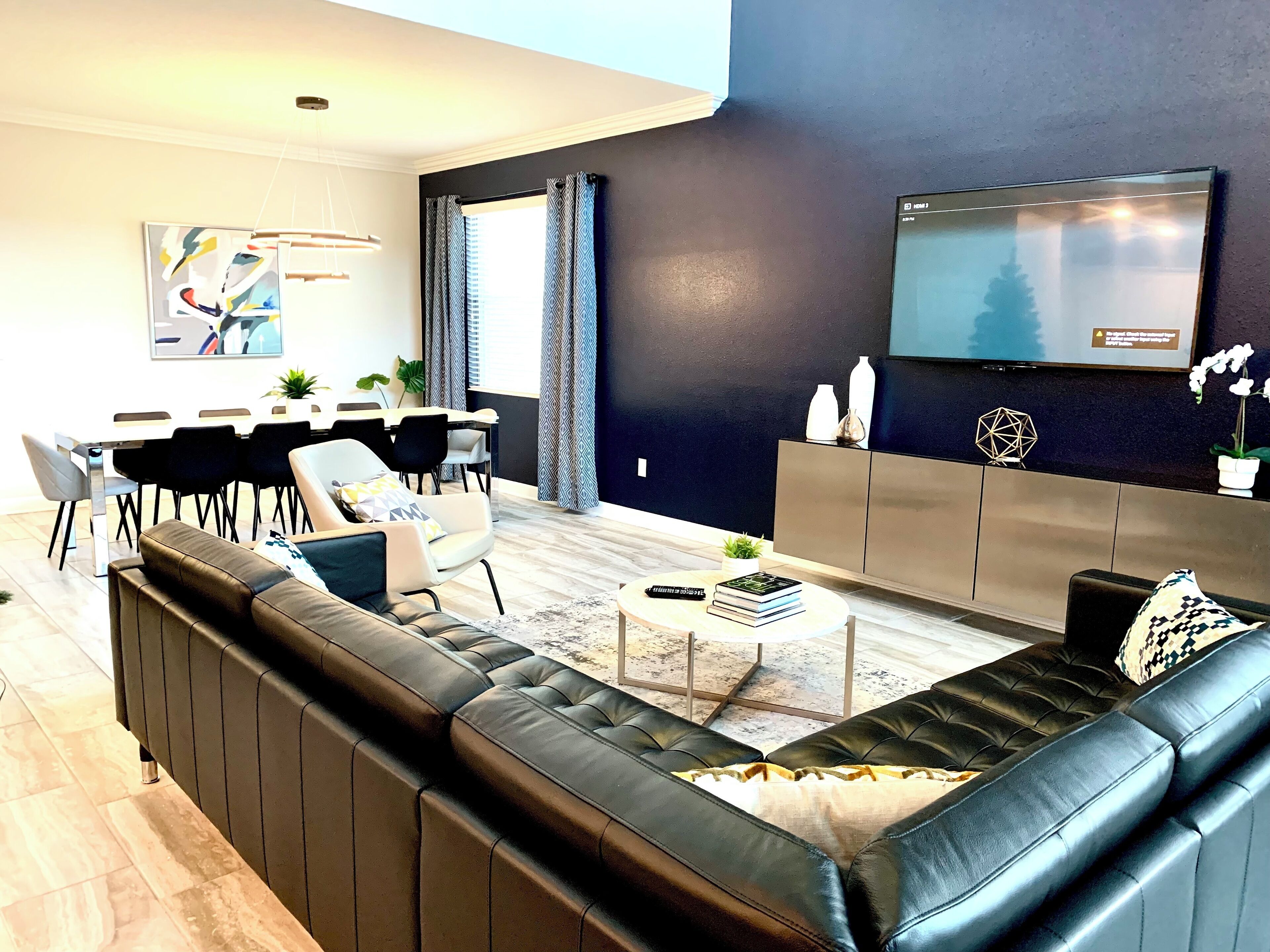 Een moderne woonkamer met een modulaire bank, een eetgedeelte met een tafel en stoelen, een flatscreen-tv aan de muur en een groot raam met gordijnen.