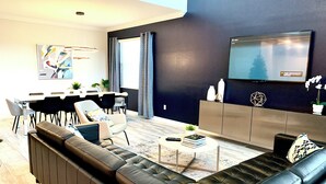A modern living room with a sectional sofa, a dining area with a table and chairs, a flat-screen TV mounted on the wall, and a large window with curtains. - Luxury 5-bedroom near Disney, Clubhouse Resort, Private Pool & Spa (Kissimmee)