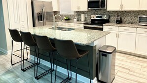 A modern kitchen with white cabinets, a granite countertop, stainless steel appliances, and a bar stool area. - Luxury 5-bedroom near Disney, Clubhouse Resort, Private Pool & Spa (Kissimmee)