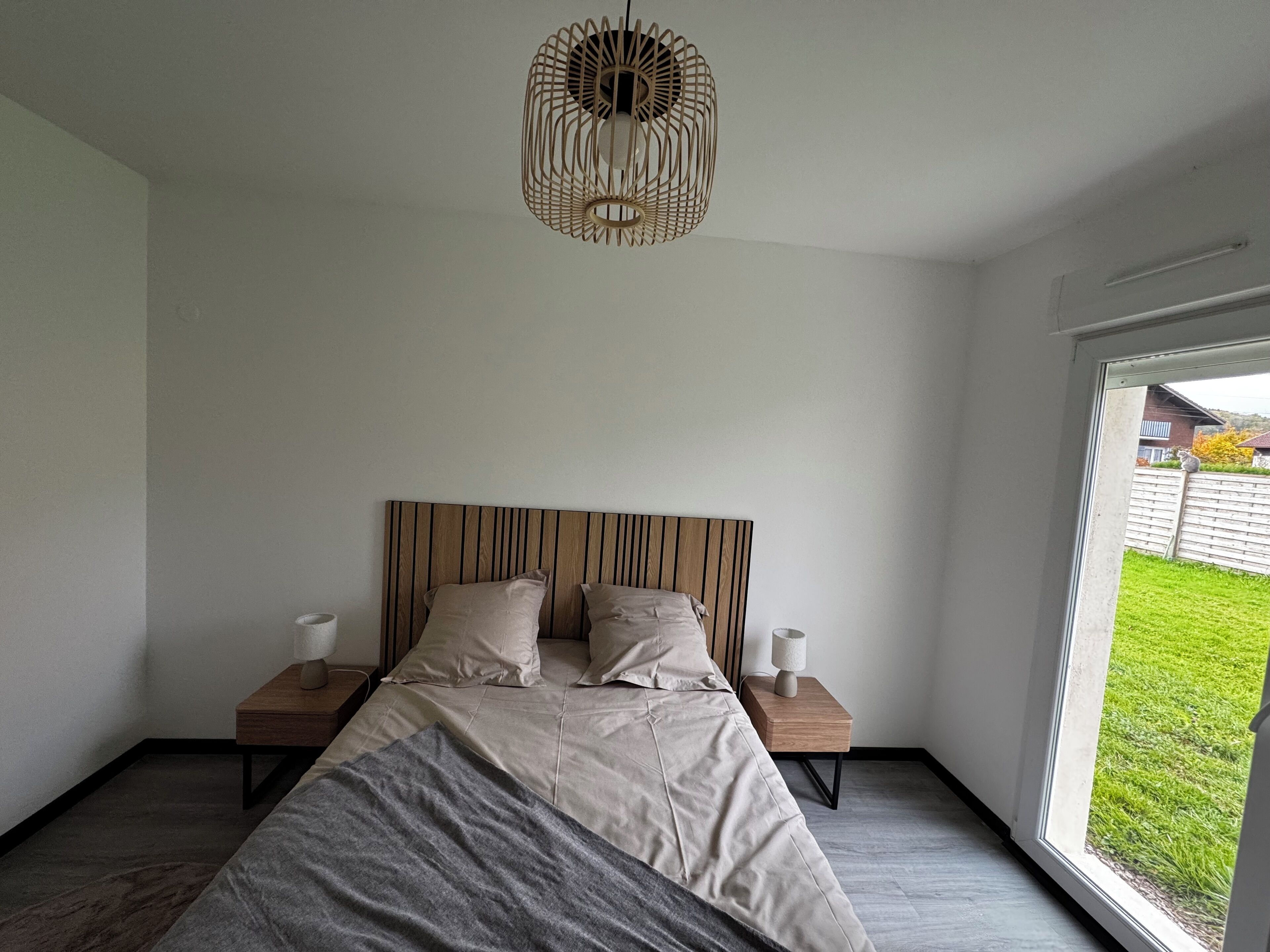 A modern bedroom with a wooden headboard, a large bed with a grey blanket, two bedside tables with lamps, and a view of a green lawn through a large window.