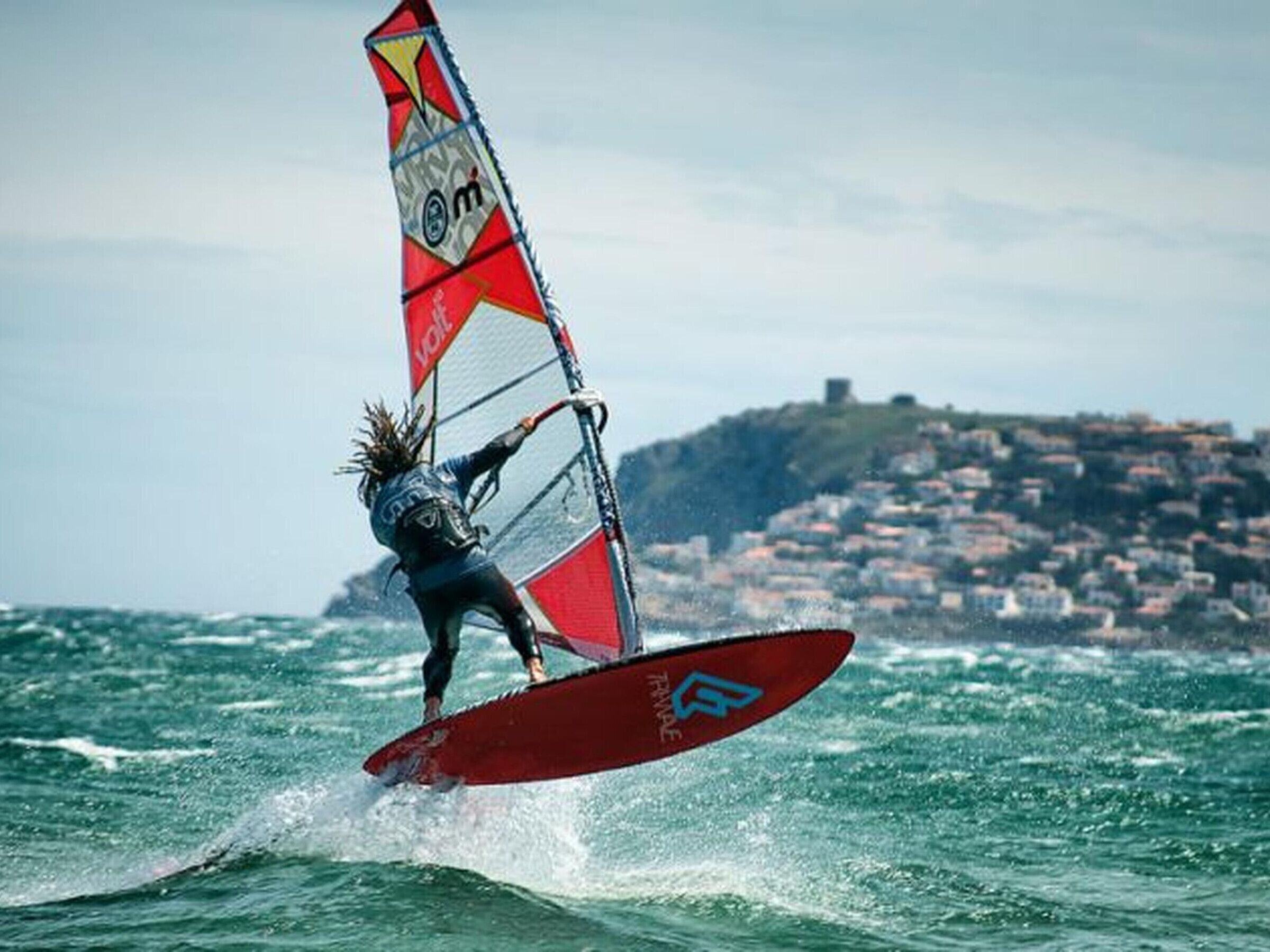 Eine Person windsurft auf einem roten Brett mit einem rot-weißen Segel, im Wasser mit einer Stadt im Hintergrund.