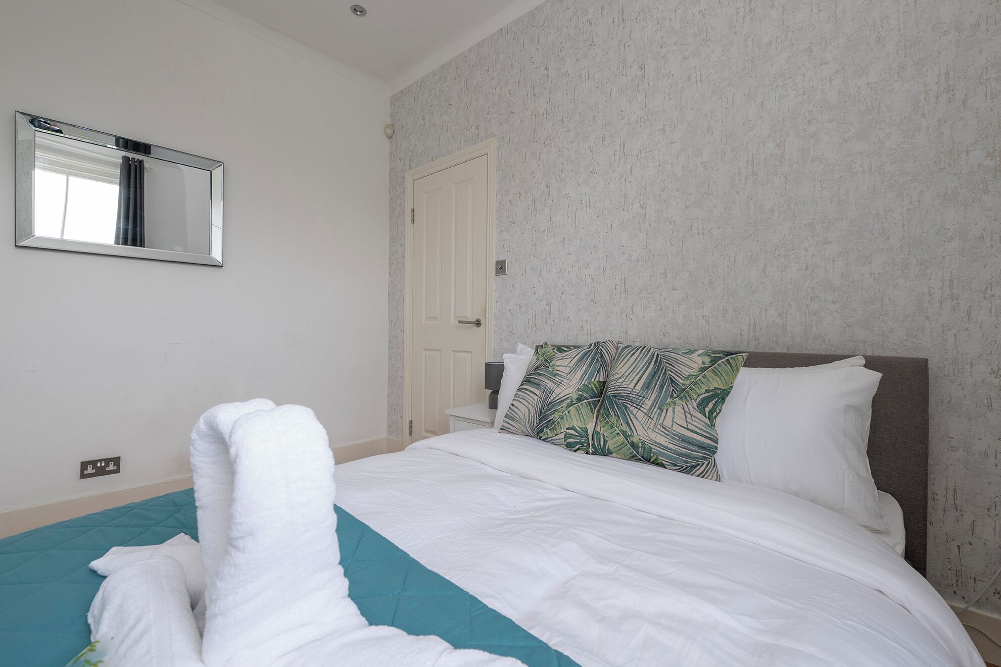 A neatly made bed with white linens and a teal accent, a large mirror on the wall, and a door with a textured wall behind it.