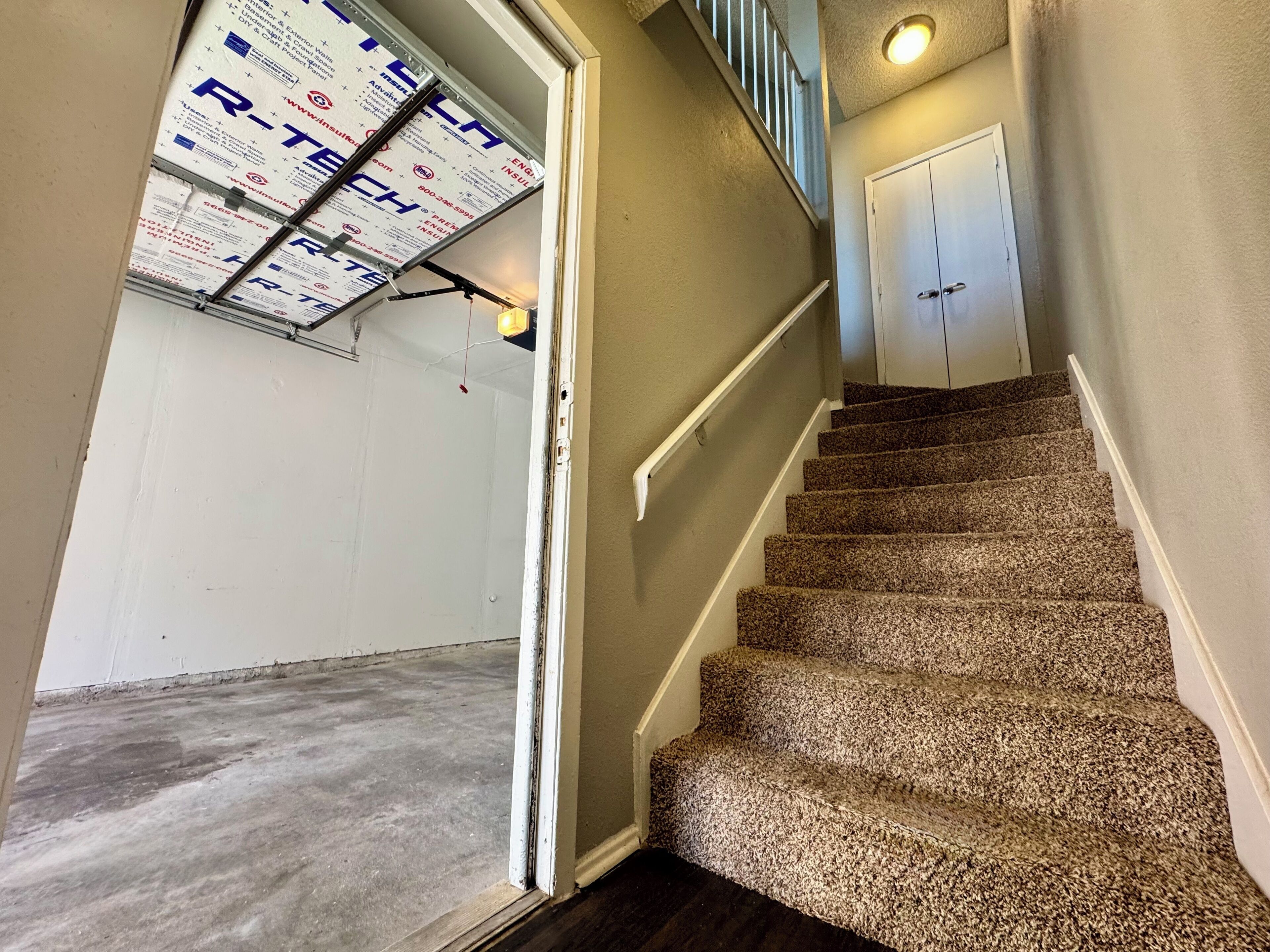 A staircase with carpeted steps leading to a door, a ceiling with insulation material, and a light fixture.