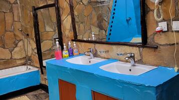 A bathroom with a stone wall, a large mirror, two sinks, a hairdryer, and a towel rack.