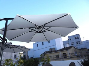 A white umbrella structure with a metal frame, set against a backdrop of buildings with white and beige facades.