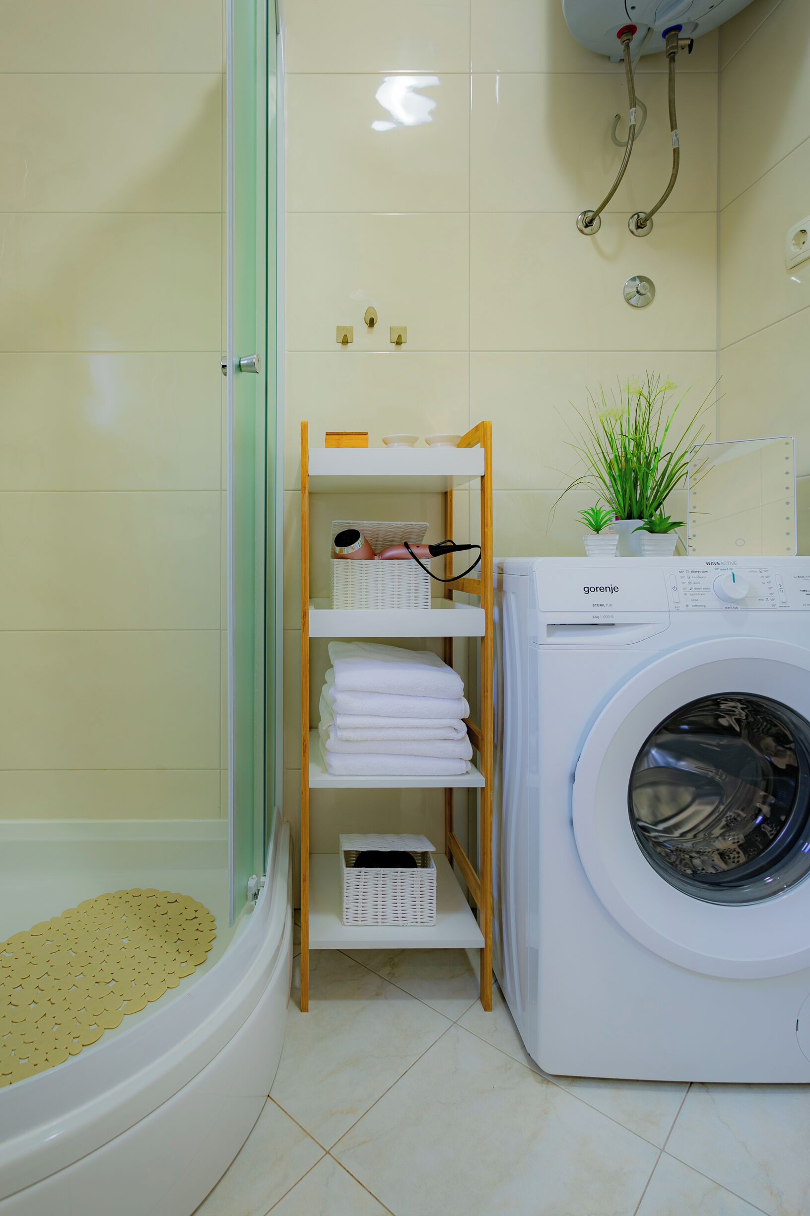 A bathroom with a glass shower enclosure, a white washing machine, and a shelf with folded towels and a potted plant.