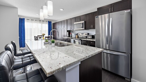 A modern kitchen with dark cabinets, stainless steel appliances, a granite countertop, and a dining area with black leather chairs. - Laurel Modern Haven (Edmonton)