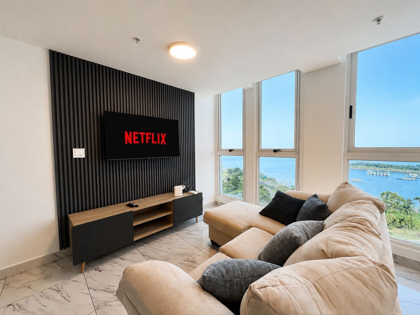 Un salon moderne doté d’un grand téléviseur à écran plat fixé au mur, d’un canapé avec des coussins, d’un meuble de rangement en bois et de grandes fenêtres offrant une vue sur la mer.