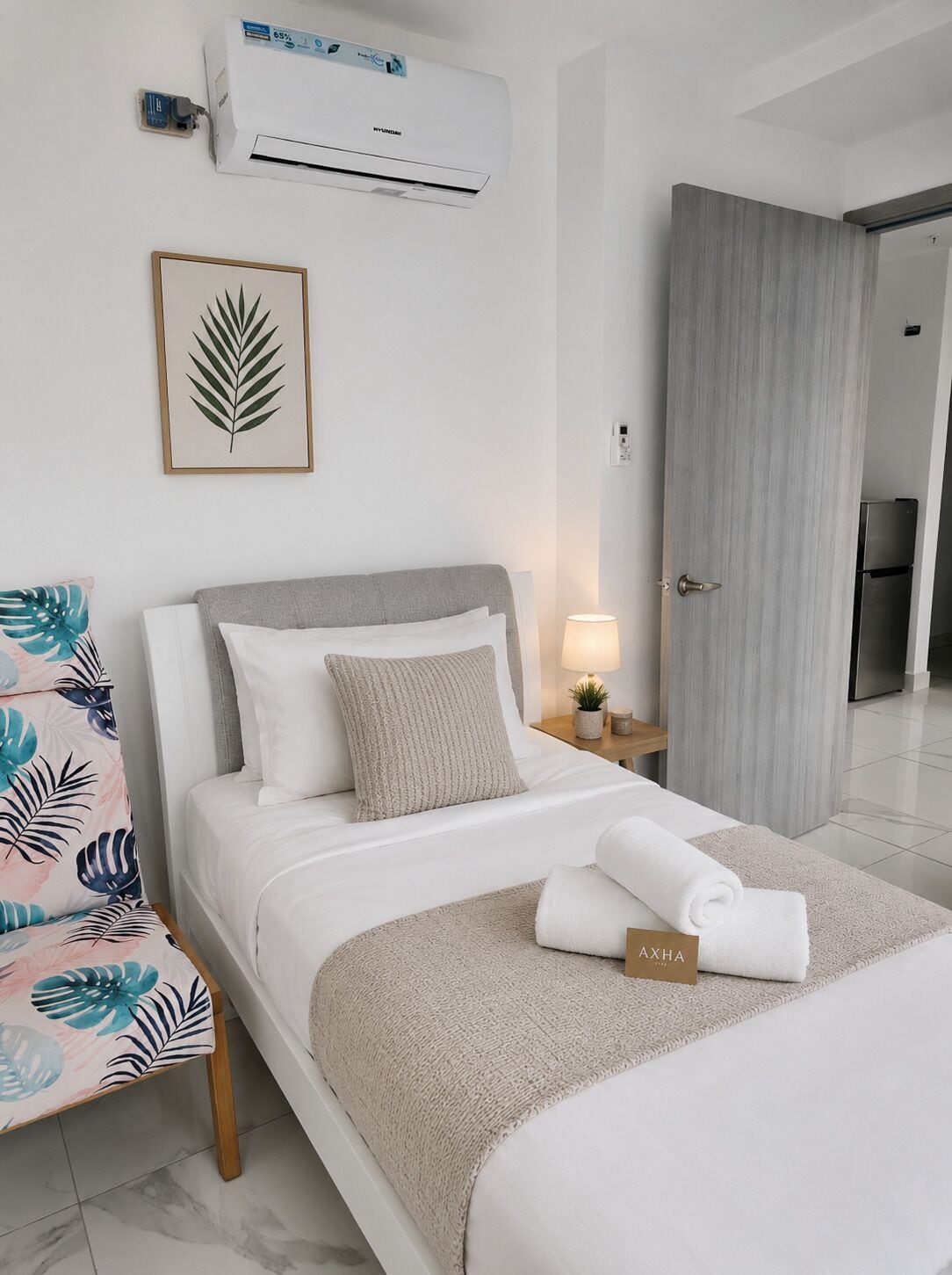 A hotel room with a bed, a bedside table with a lamp, an air conditioner, and a framed leaf artwork on the wall.