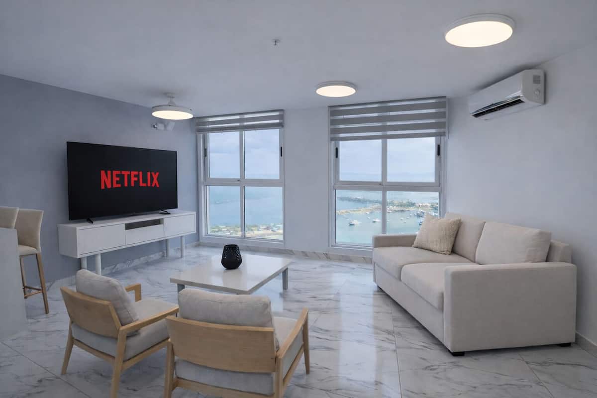 Una sala de estar moderna con un televisor de pantalla plana, un sofá, dos sillones, una mesa de centro y ventanas amplias con vista al mar.