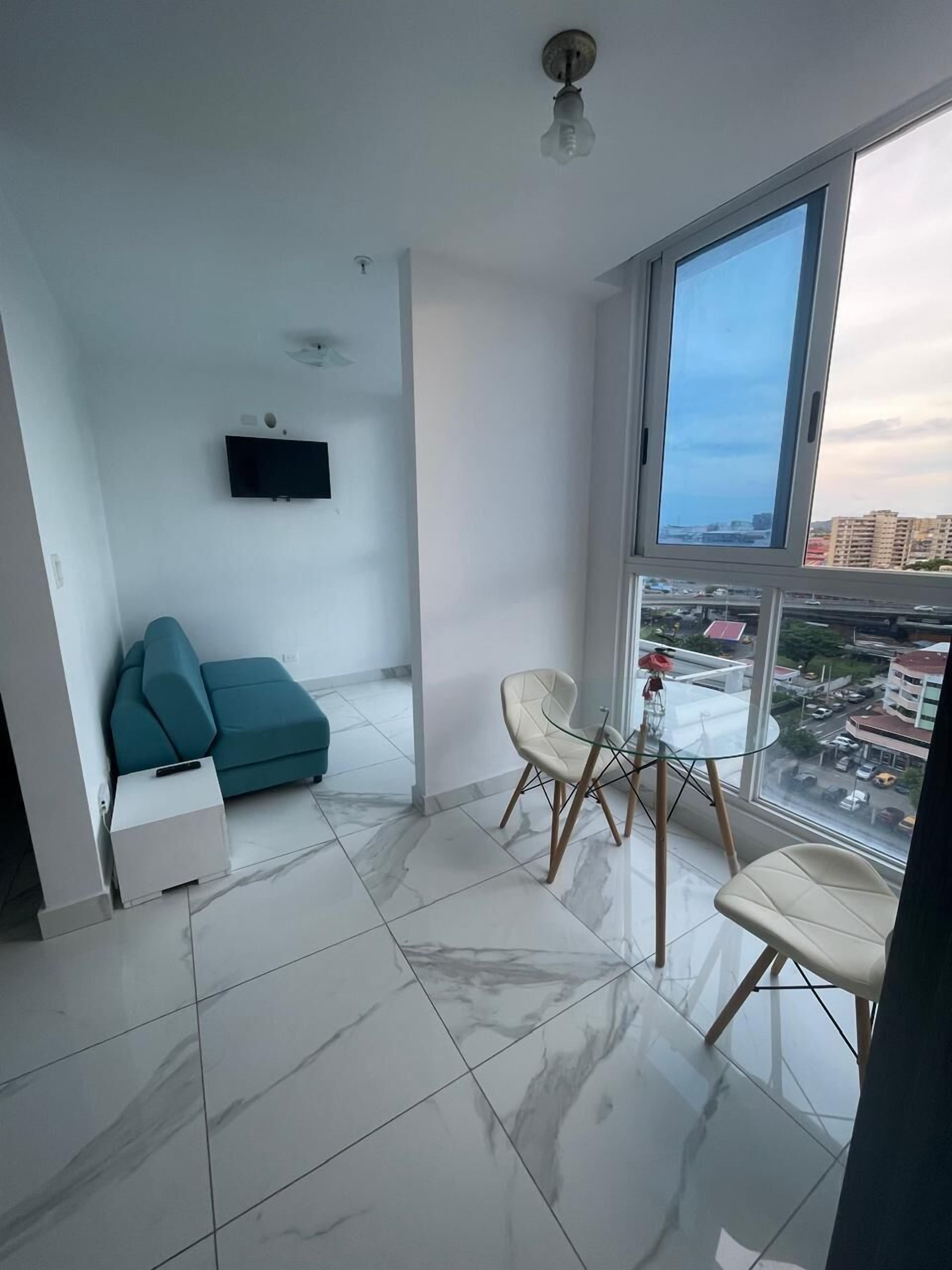 A modern living room with a teal sofa, a glass table with chairs, a mounted TV, and a large window offering a city view.