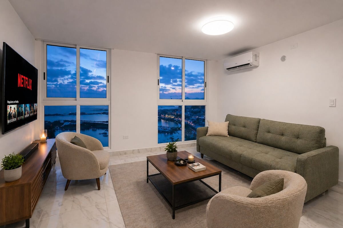 Una sala de estar moderna con un televisor grande, asientos cómodos, una mesa de centro y vistas a un paisaje urbano a través de grandes ventanales.